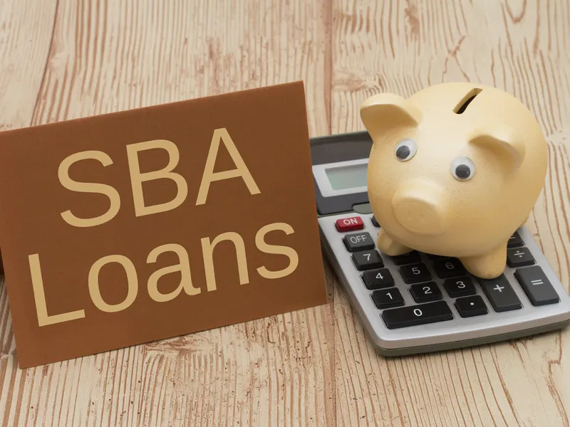 Brown card reading SBA Loans beside a gold piggy bank sitting on a calculator, on a light wooden table
